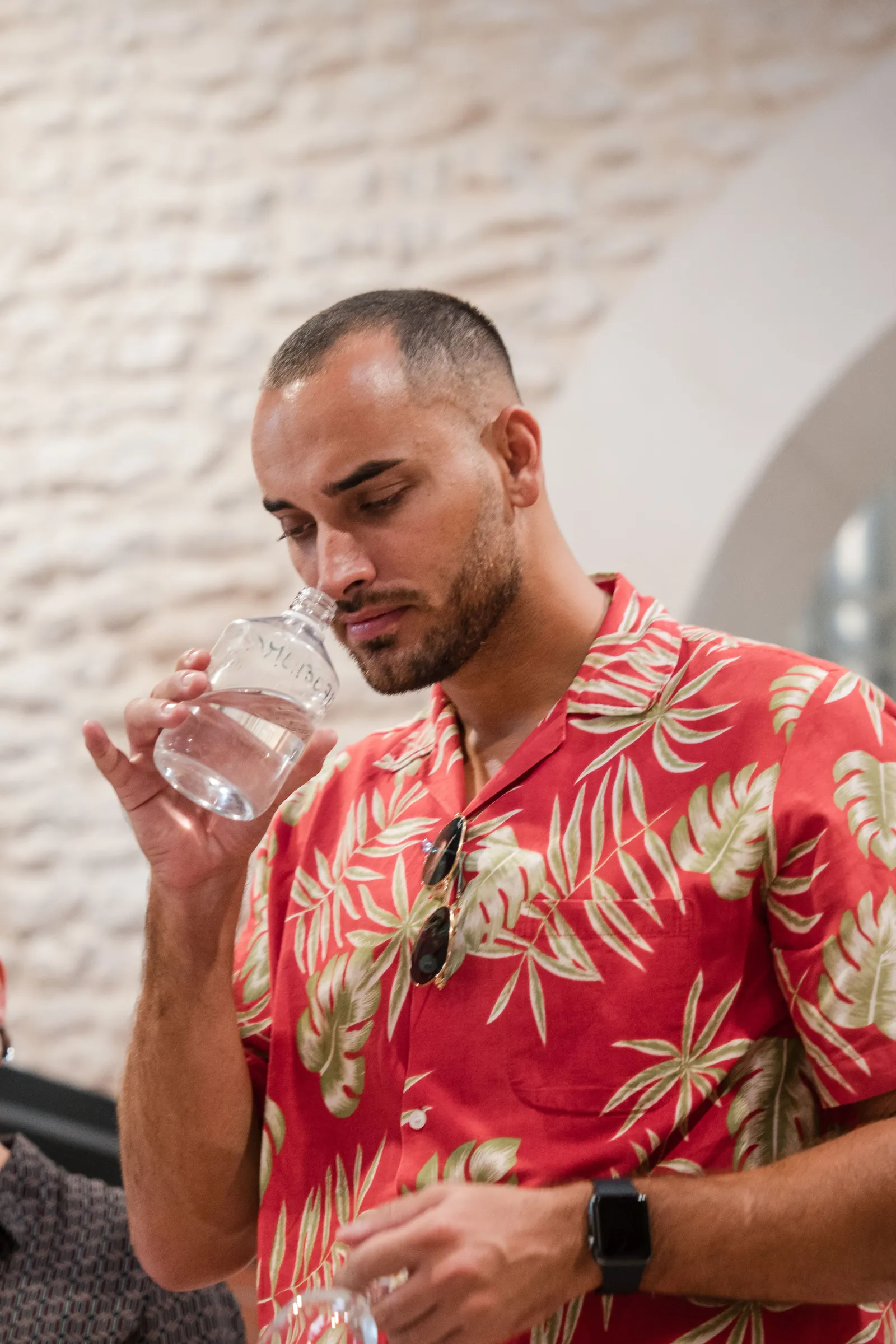 A man wearing a red floral patterned shirt drinks a glass of clear water at Citadelle Gin Distillery in Bordeaux, standing in front of a white brick wall.