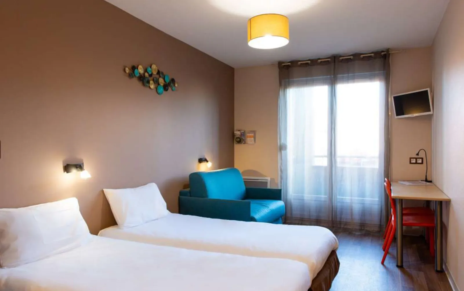 Youth hostel room featuring two single white beds, blue sofa, desk, balcony access, turquoise and gold wall decoration, yellow ceiling light