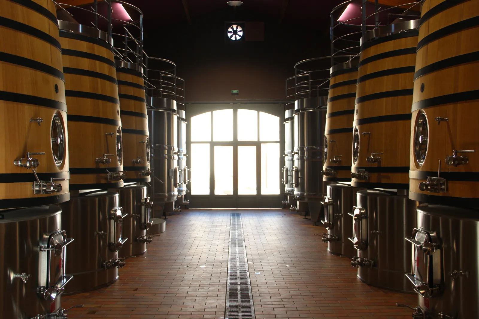 Interior of a winery with wooden fermentation barrels banded with metal, symmetrically arranged on either side of a central aisle, with a glass door visible at the end.