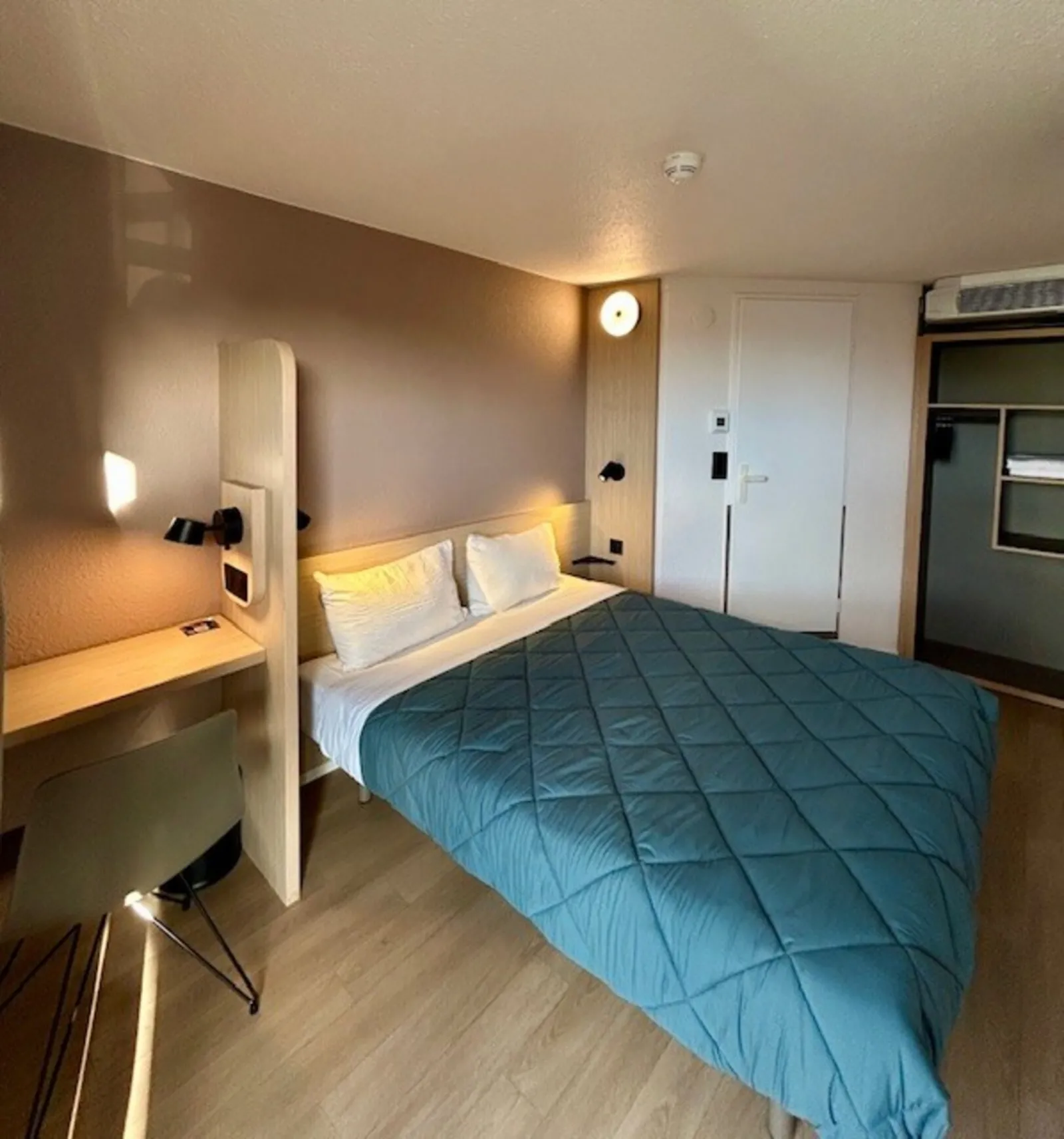 Modern hotel room featuring a double bed with blue-grey quilted comforter, integrated desk with warm lighting, white wardrobe, and kitchenette visible in background