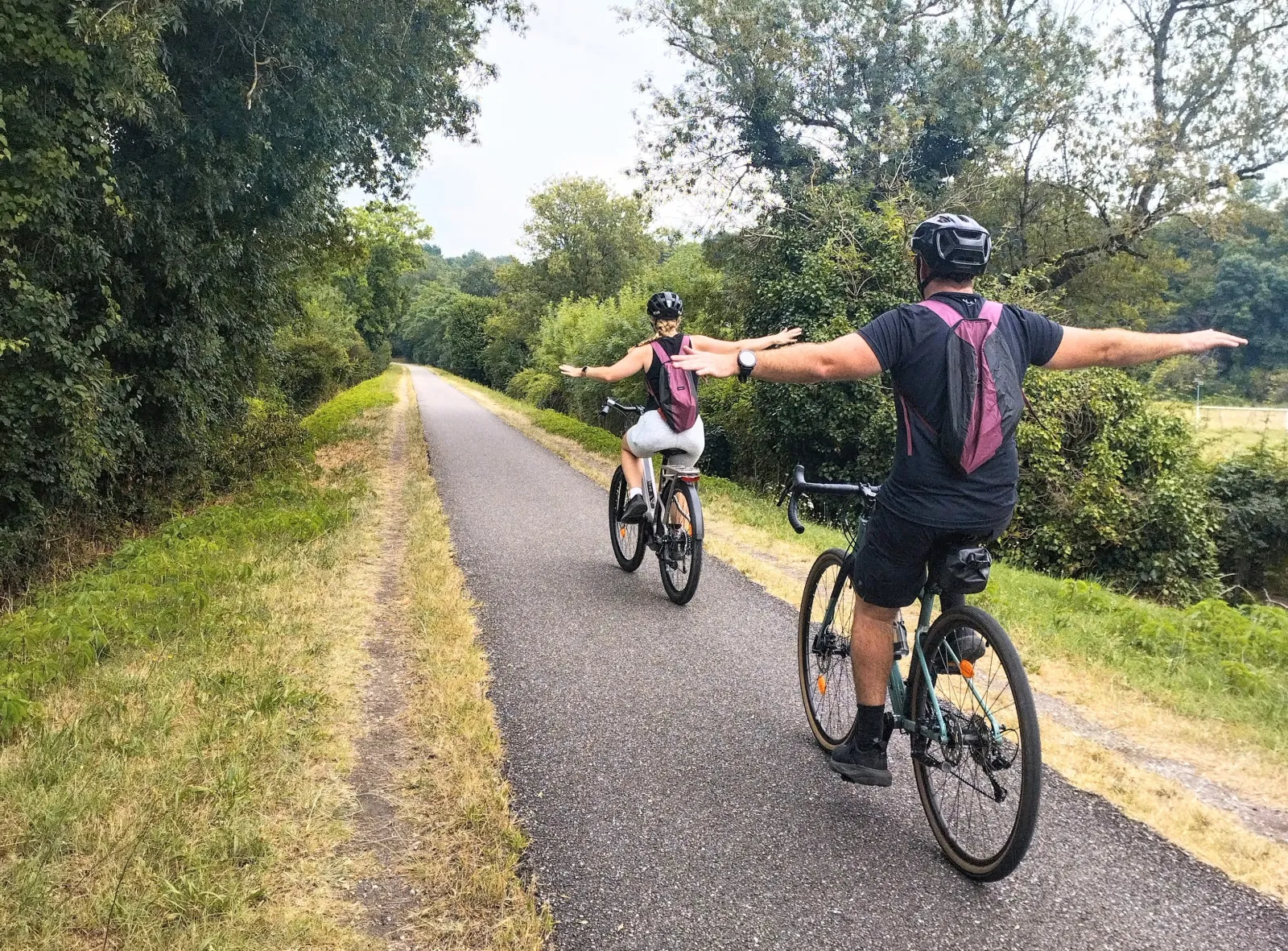 Two gravel bike riders cycling on a tree-lined path through Bordeaux vineyards, with one rider raising their arms in celebration.