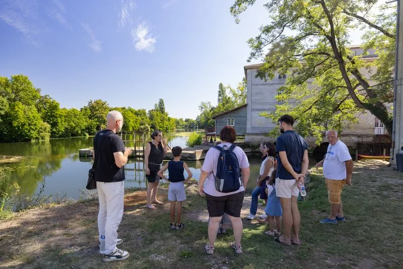 A group of tourists and visitors gather near the Moulin de Porchères in Bordeaux, by the riverside, with a historic building in the background and lush green trees under a sunny sky.