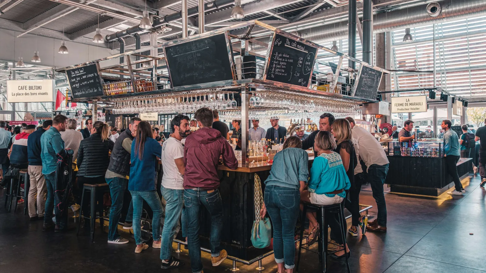 Interior view of Halles de Bacalan in Bordeaux showing a crowd of visitors gathered around a wine bar. Suspended glass shelves and menu chalkboards are visible above the counter in this modern covered market.