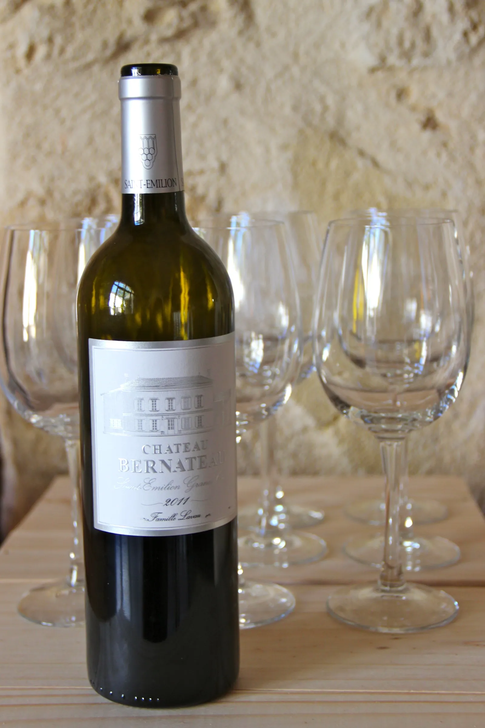 A bottle of Château Bernateau red wine from Bordeaux surrounded by empty wine glasses on a wooden table against a stone wall.