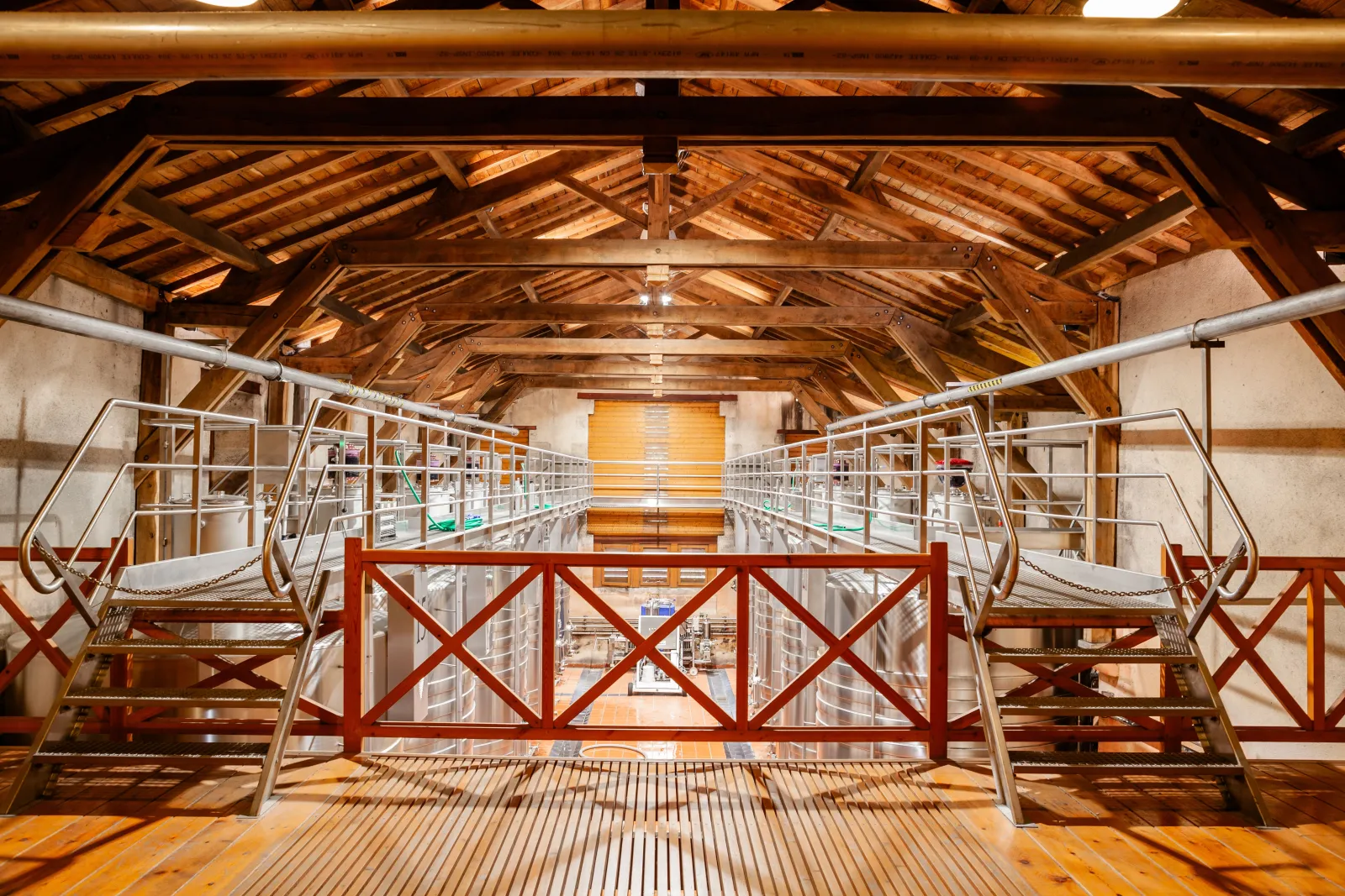 Interior view of a wooden mezzanine at Château Desmirail in Bordeaux, featuring exposed wooden beams, red and white metal staircases, mesh railings, and storage units along the walls.