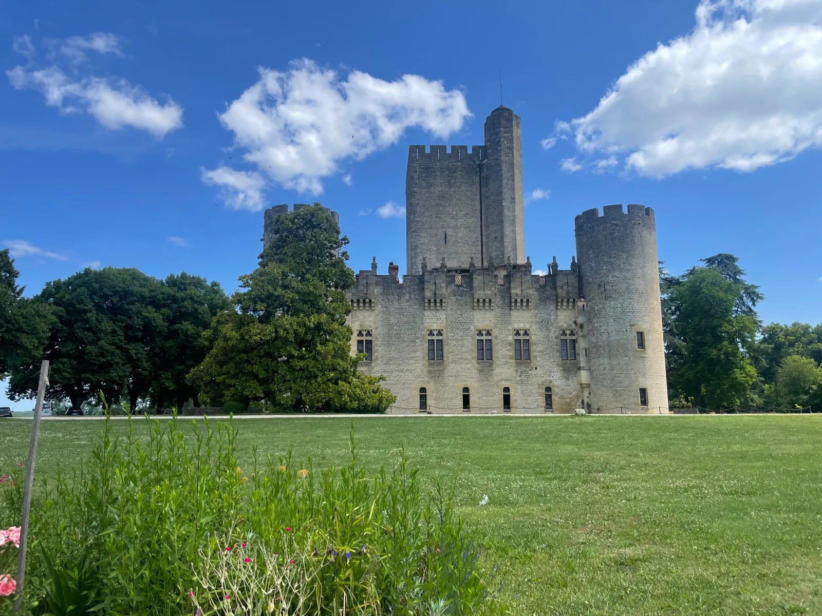 Roquetaillade Castle, a medieval stone fortress with crenellated towers, surrounded by lush green grounds under a clear blue sky.