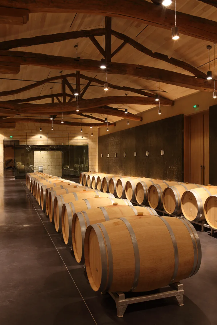 Wine aging cellar at Château Soutard in Bordeaux featuring rows of oak barrels beneath exposed beam ceilings with contemporary lighting