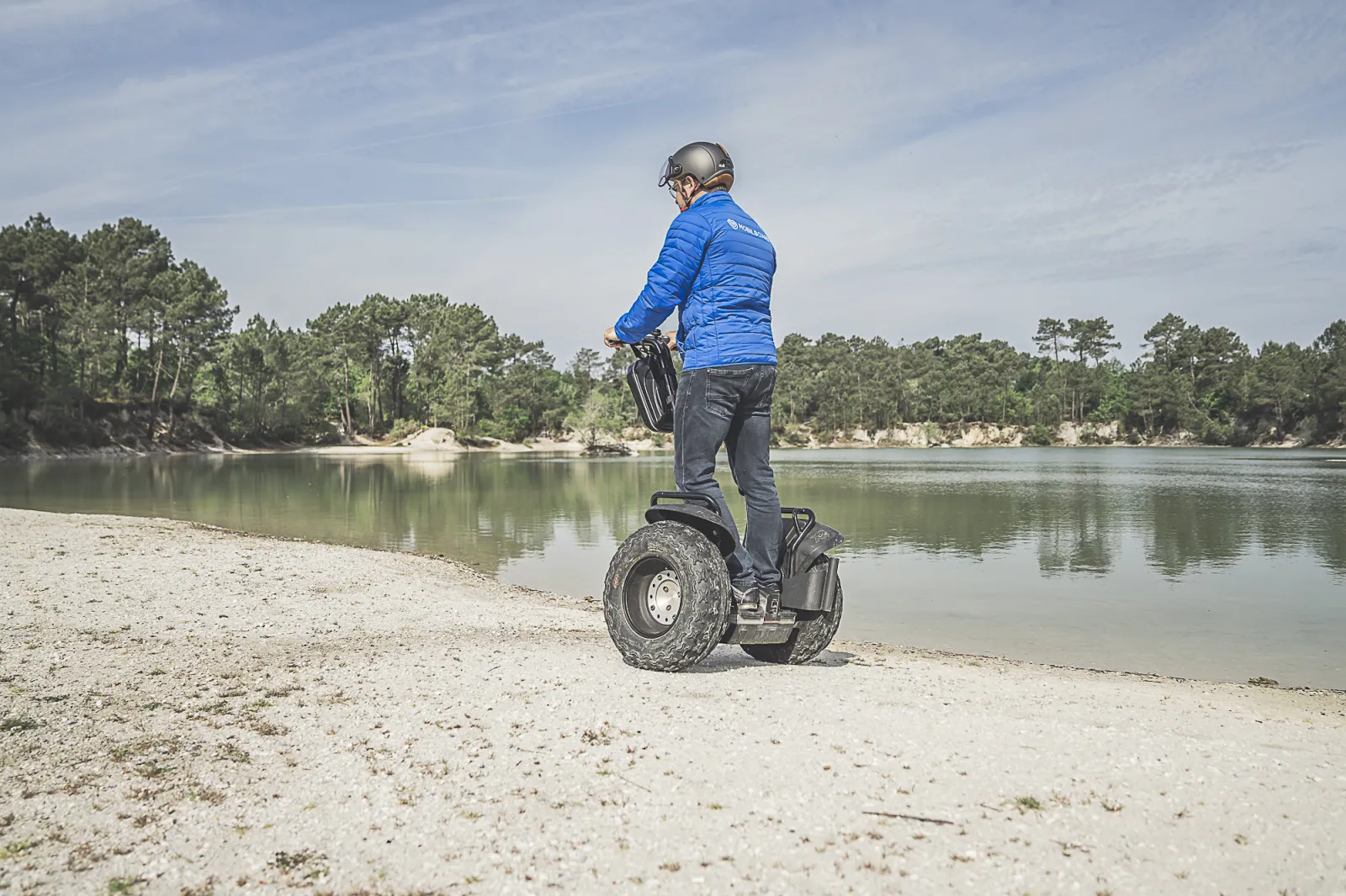 Person in blue jacket and helmet riding a Segway on a sandy beach overlooking a calm river lined with trees, Bordeaux area