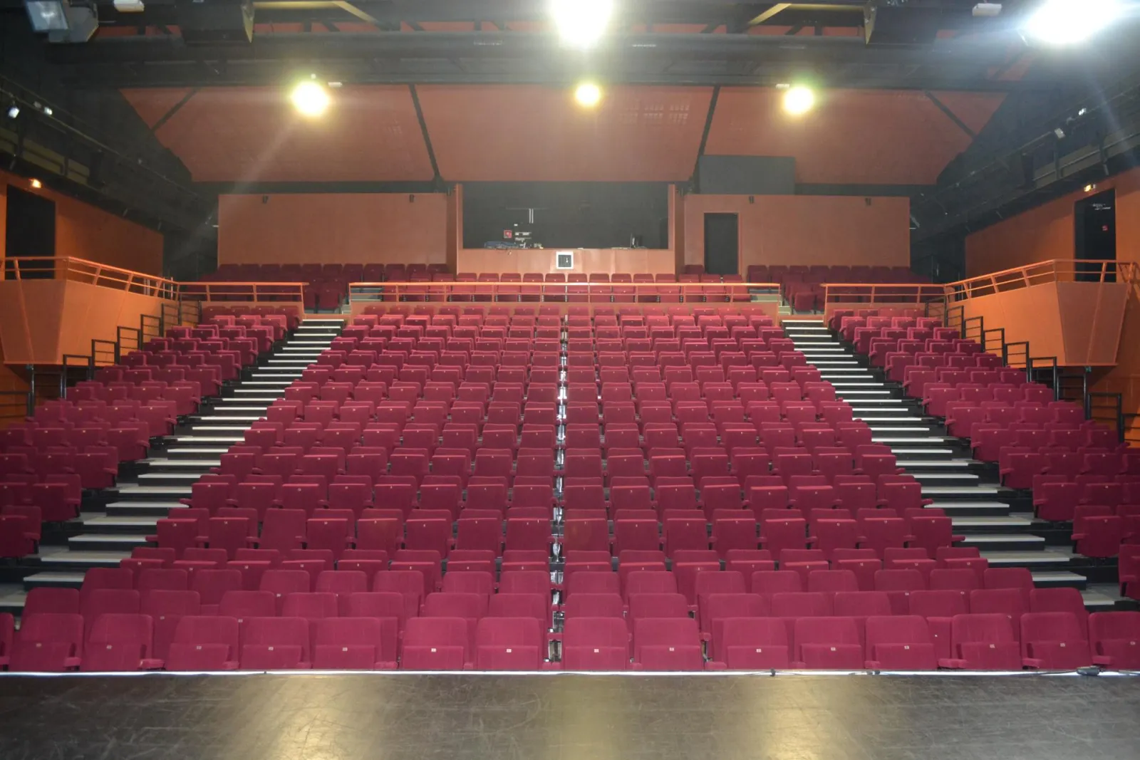 Empty cinema auditorium with red seats arranged in rows, gray emergency stairs, orange walls, gray ceiling with lighting, and black screen at the back, at Espace Culturel Treulon in Bordeaux.