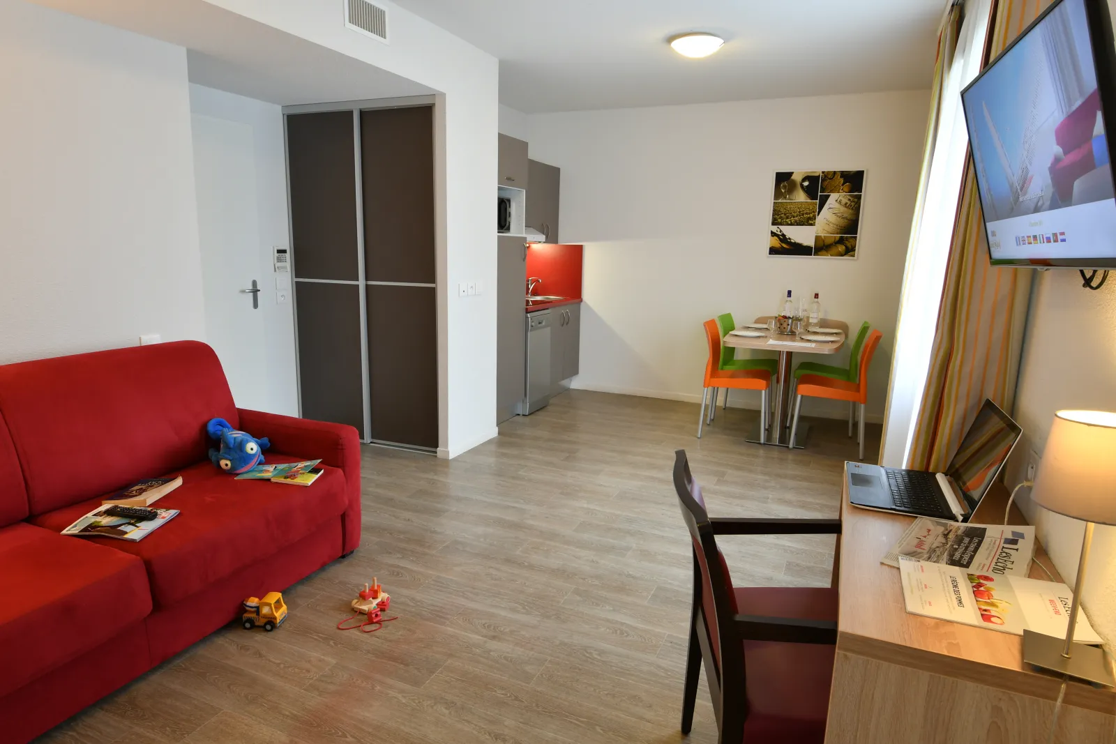 Modern living space in a Bordeaux apartment-hotel featuring a red sofa, equipped kitchenette, dining table with colorful chairs, work desk with laptop, and wall-mounted television. Children's toys scattered on the floor.