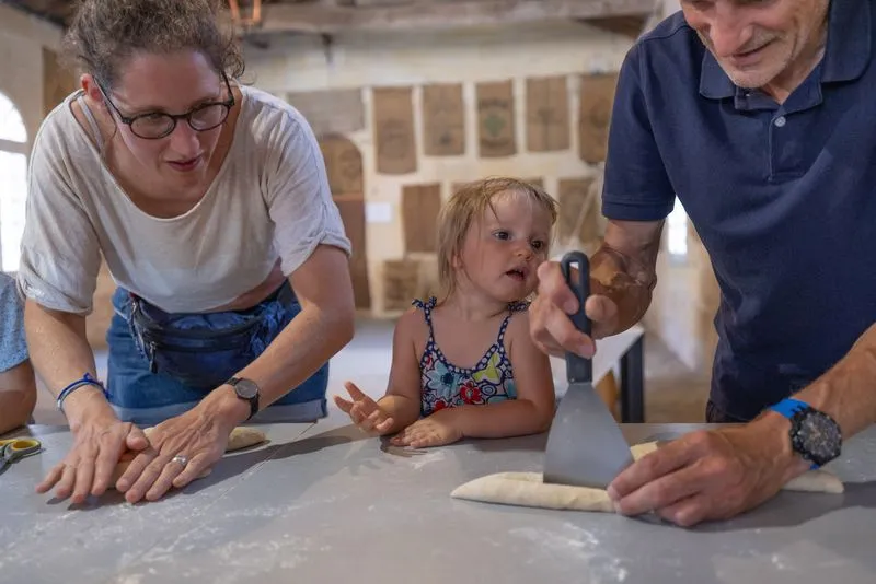A family discovers traditional bread-making at the Moulin de Porchères in Bordeaux. A young girl watches intently as an adult shapes dough on a floured work surface, surrounded by two other people.