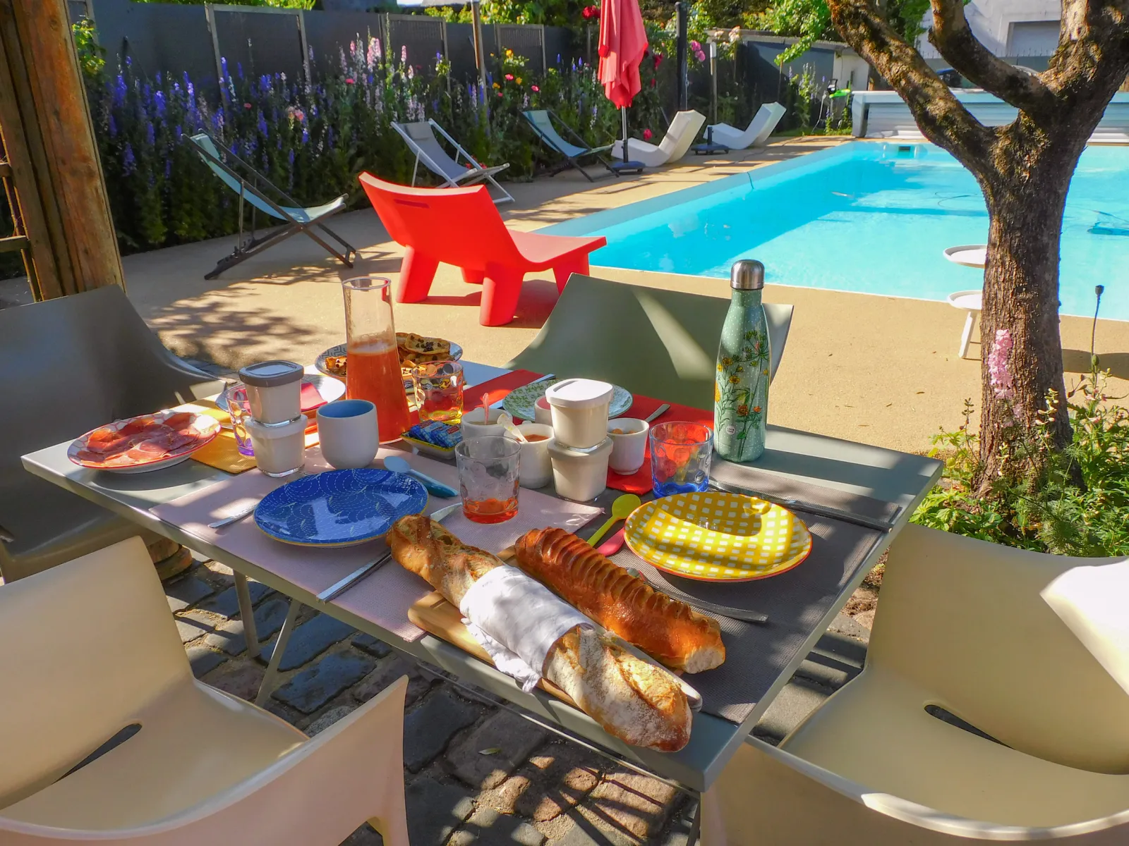 Breakfast table set poolside with fresh bread, fruits, drinks and colorful tableware, in a sunny garden with red and blue loungers at Gatto Verde, Bordeaux.