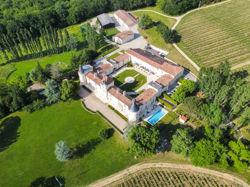 Aerial view of Château Toulouse-Lautrec near Bordeaux, featuring a white manor with towers surrounded by vineyards, green fields and forests, with a swimming pool and farm buildings