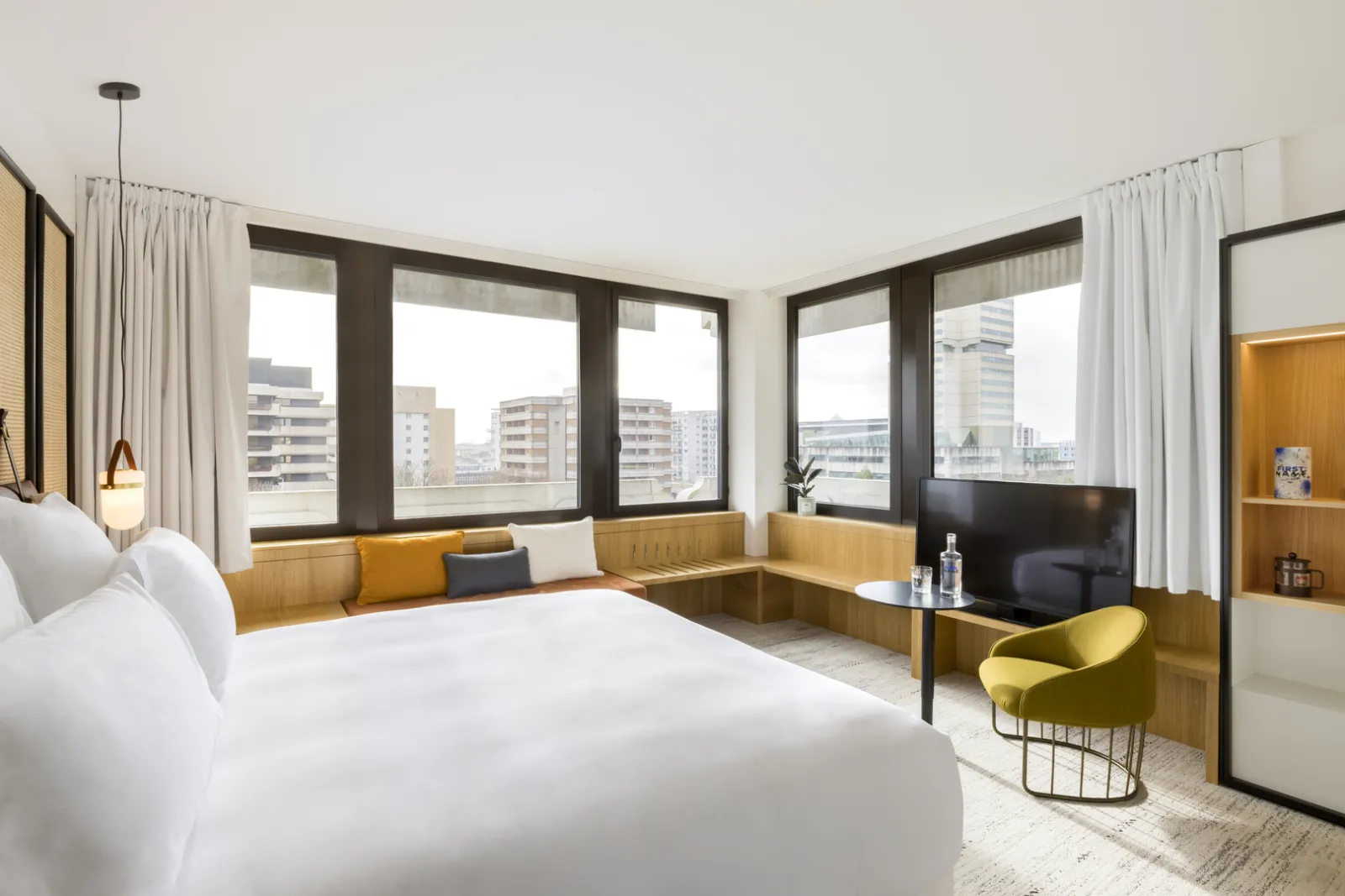 Spacious luxury hotel room in Bordeaux featuring a white bed, mustard-yellow seating area, flat-screen TV, and panoramic windows overlooking the city skyline.