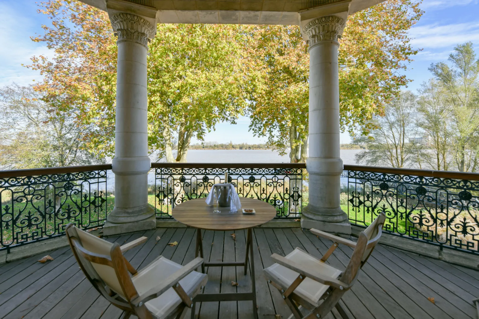 Covered terrace at Château Grattequina in Bordeaux featuring two Corinthian columns, wrought iron railing, two lounge chairs, overlooking a lake with tree-lined avenue in autumn