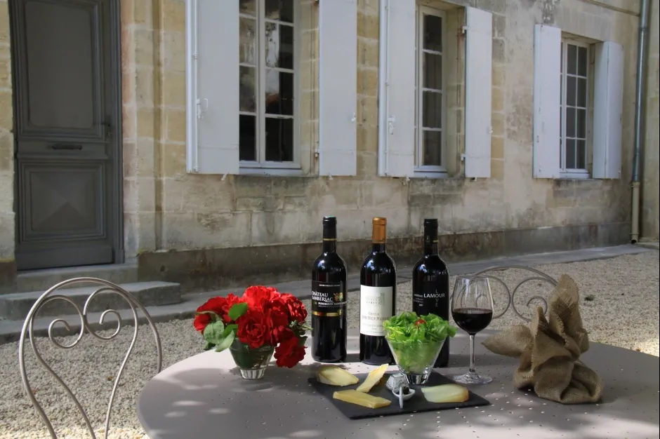 Outdoor table setting with three bottles of Bordeaux wine, glasses, red flowers, cheese and bread displayed in front of a stone building with white shutters