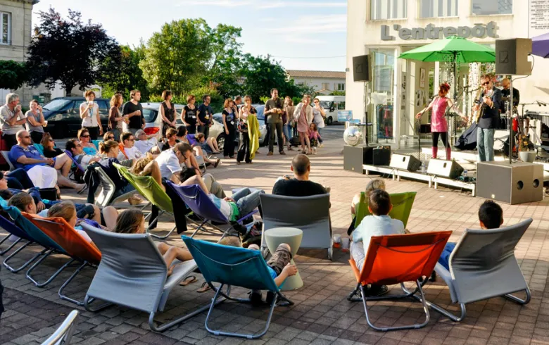 Outdoor community event at Entrepôt du Haillan in Bordeaux featuring people seated in colorful deck chairs and standing visitors, with a green umbrella, in a festive and welcoming atmosphere.