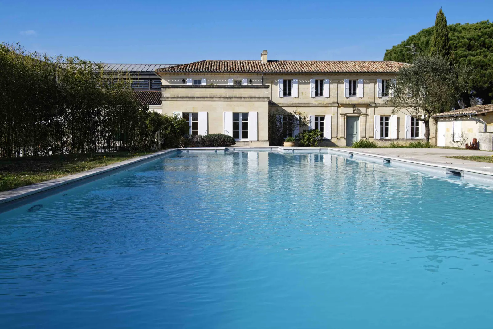 A turquoise rectangular swimming pool overlooking a classic Bordeaux mansion with blue shutters, surrounded by lush greenery under a clear blue sky.