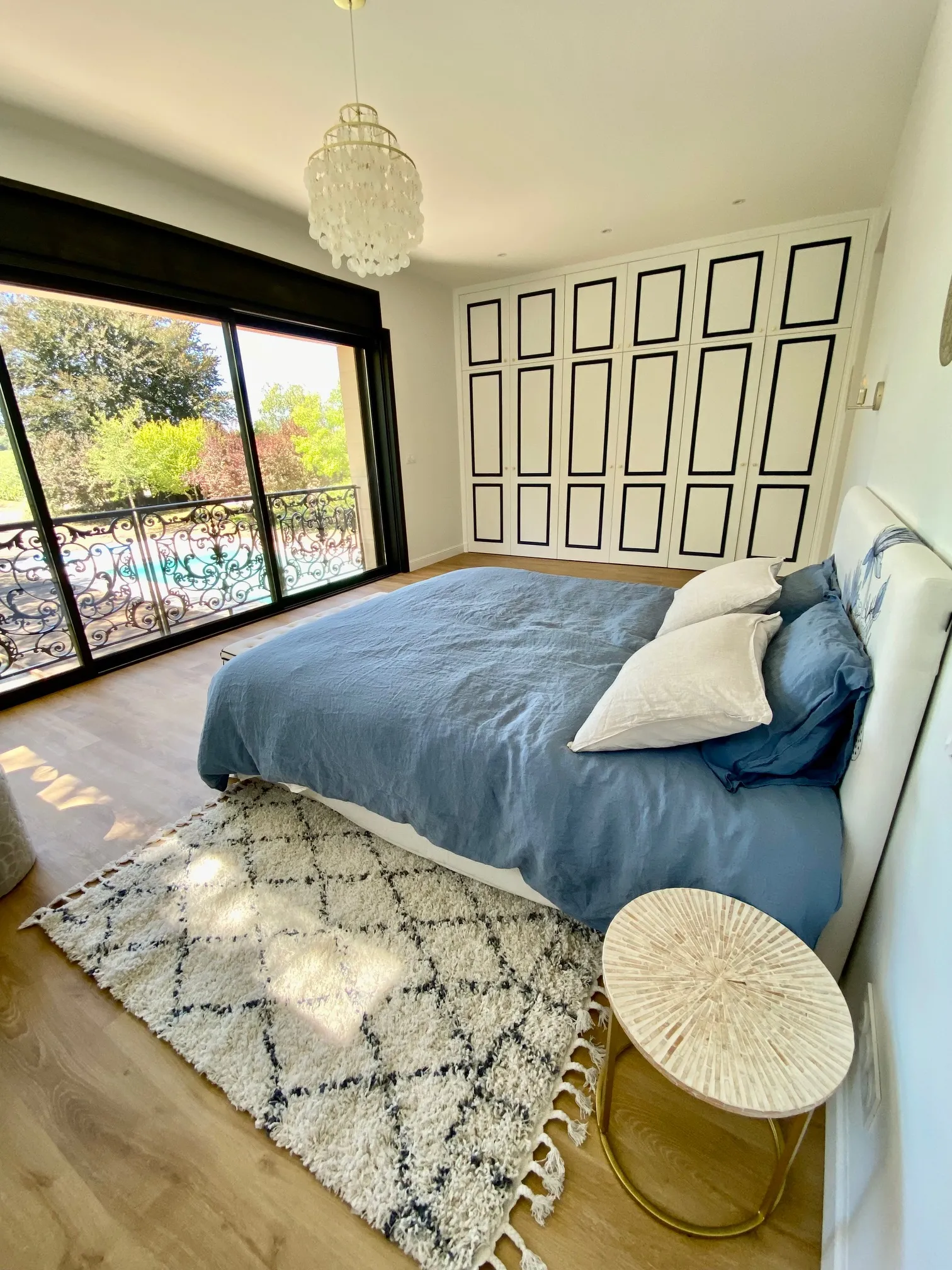 Modern bedroom with blue bed, glass doors opening onto a balcony, white paneled wardrobes and crystal chandelier at Maison L'Eperon in Bordeaux