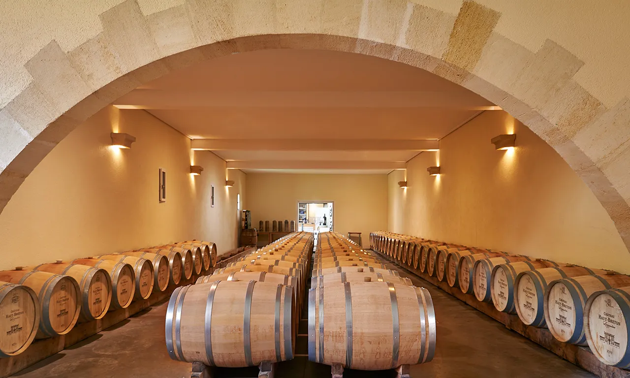 Wine aging cellar at Château Haut Breton Larigaudière in Bordeaux, featuring rows of oak barrels lined under a stone barrel vault with warm ambient lighting.
