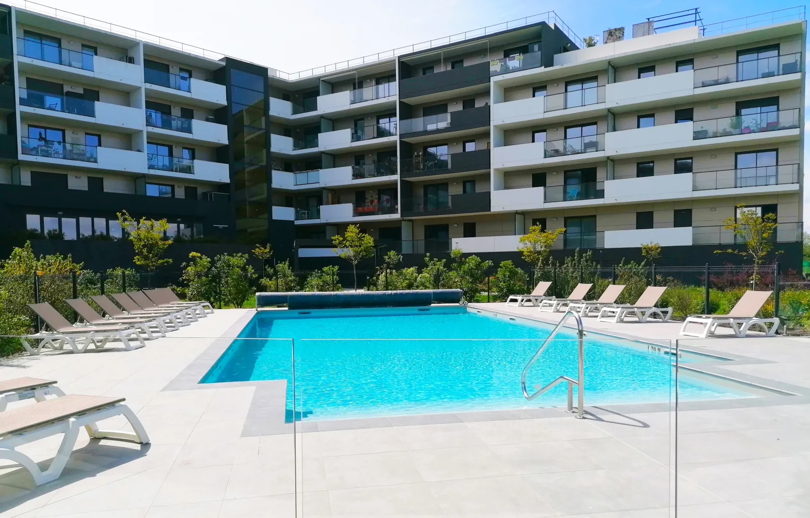 Outdoor swimming pool with turquoise water surrounded by white loungers, facing modern contemporary apartment buildings in Bordeaux Bruges.