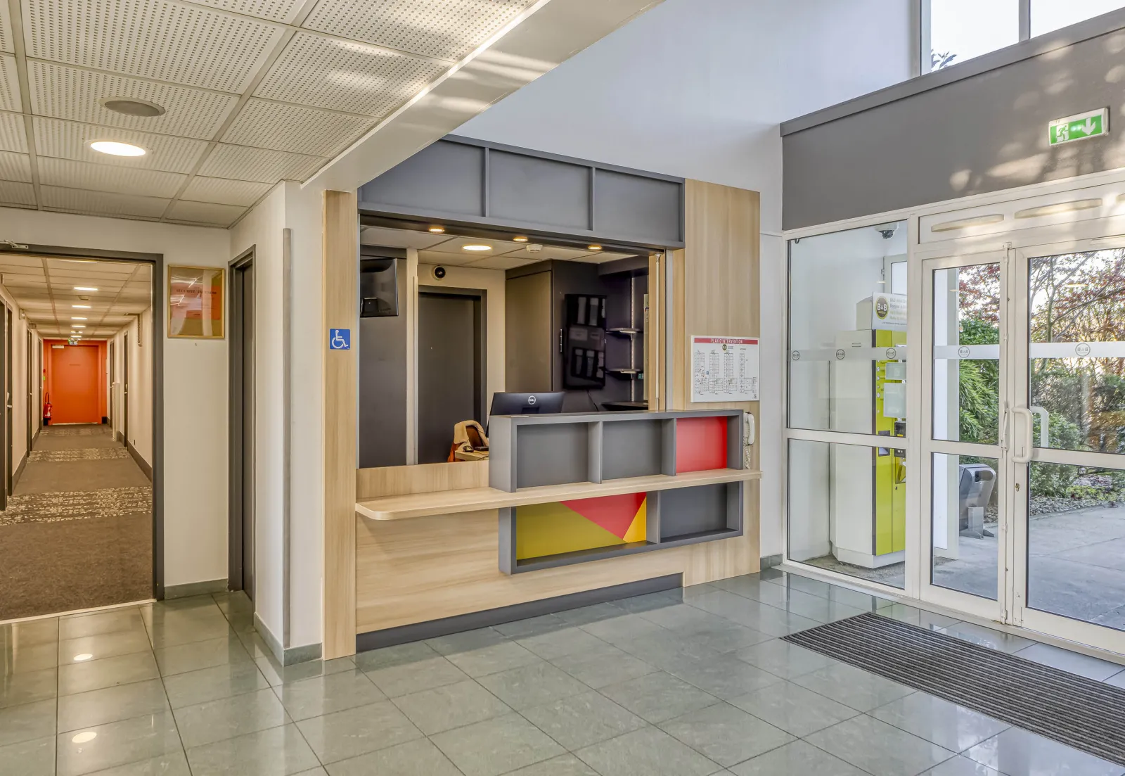 Modern reception hall of B&B Hotel Bordeaux Bruges featuring light wood desk, colorful shelving, glass doors, access corridor and accessibility signage