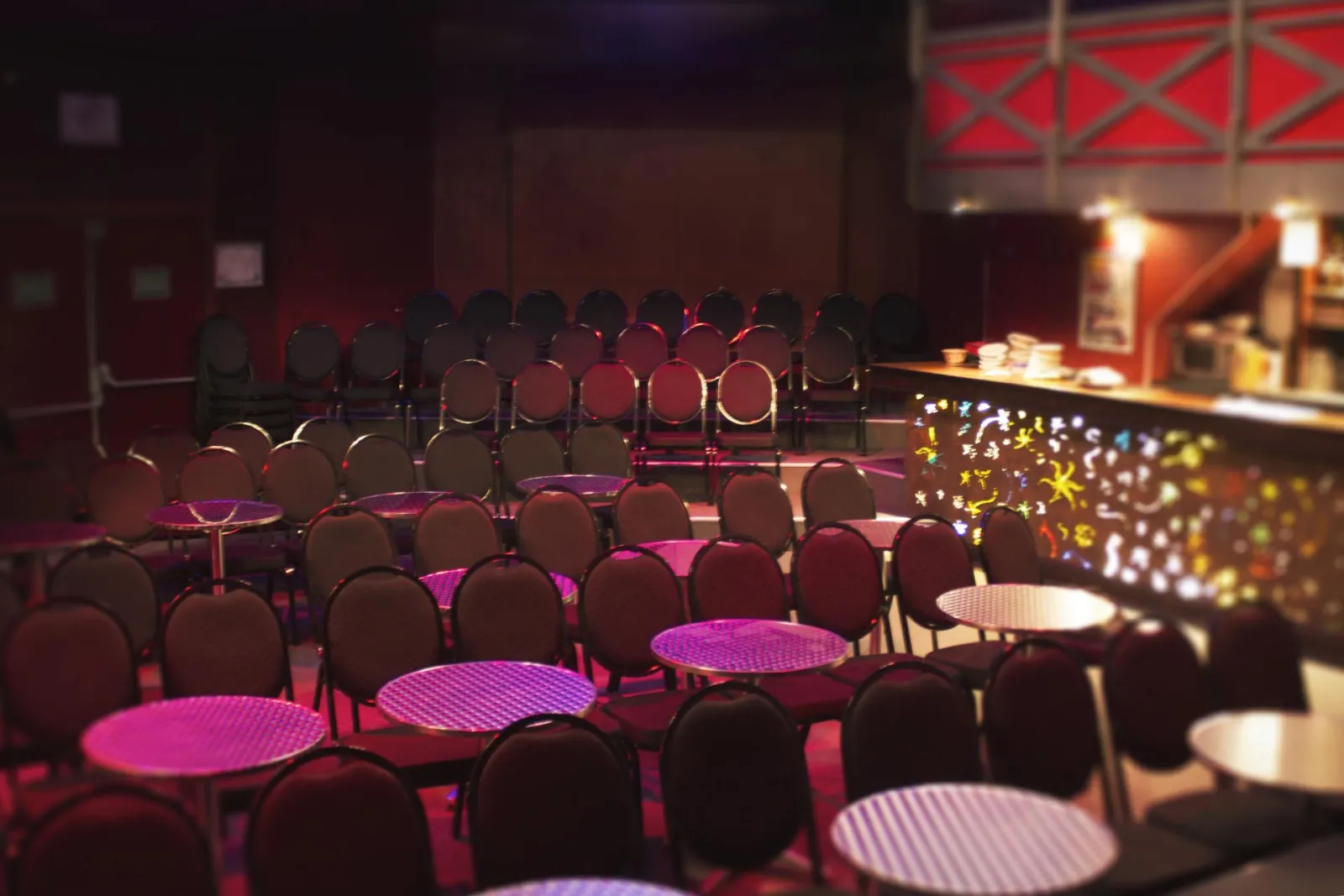 Theater auditorium at the Beaux Arts Theatre in Bordeaux featuring black chairs arranged facing a stage lit in pink and purple, with high tables in the foreground.