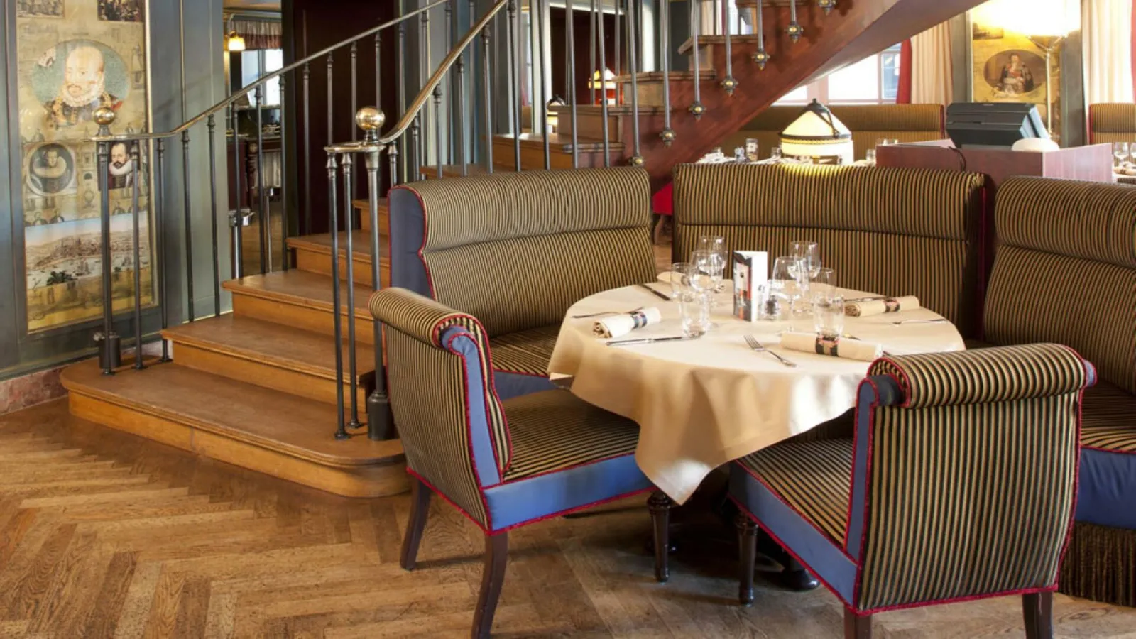 Elegant dining room of Le Bordeaux restaurant featuring striped blue and gold armchairs, a set round table, wooden staircase, and historical displays on the walls.
