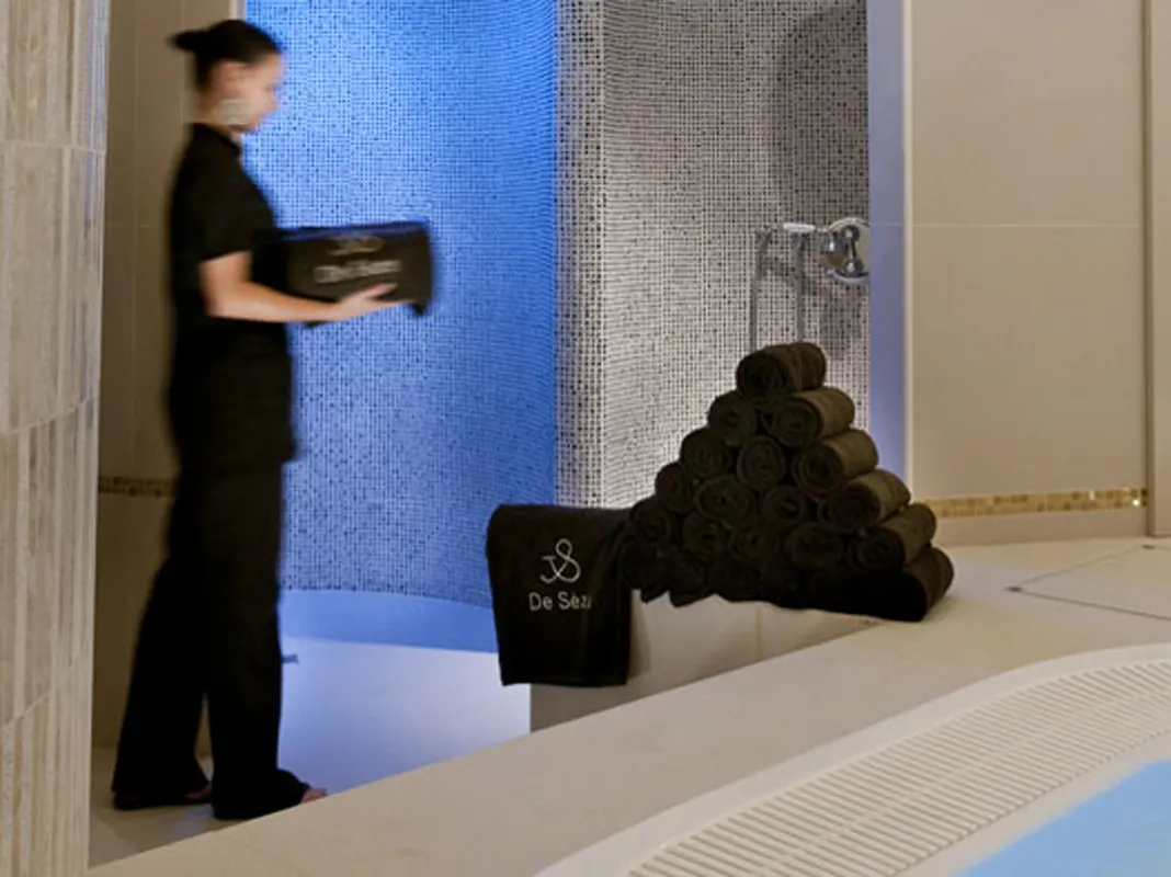 Interior of the Hôtel de Sèze spa in Bordeaux featuring a staff member in black attire near a blue water basin, stacked black towels, and textured tile panels in the background.