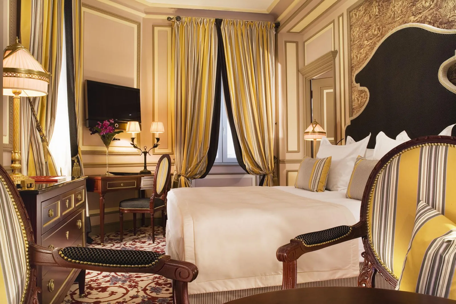 Luxurious bedroom at Intercontinental Grand Hotel Bordeaux featuring a king-size bed, ornate wooden furniture, gold and black striped curtains, wall-mounted TV, and classical elegant decor.