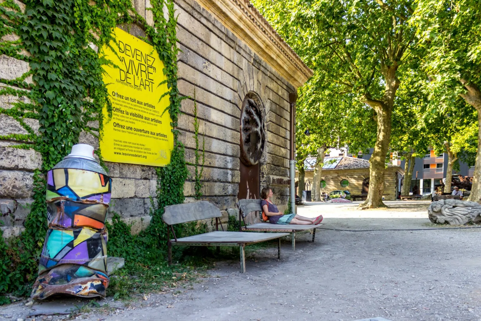 Stone courtyard with ivy-covered historic buildings, yellow « Become art supplies » poster, colorful sculpture, public benches and person sitting, trees in background