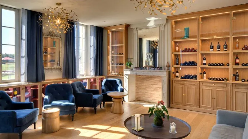 Sophisticated wine tasting room at Château Fage in Bordeaux featuring navy blue armchairs, glass-fronted wooden wine cabinet, marble bar counter, golden chandelier and vineyard views