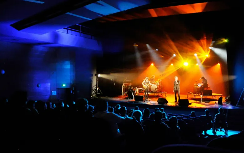 Live concert at Entrepôt du Haillan in Bordeaux featuring a stage lit in orange and yellow, musicians performing, and an audience illuminated by blue stage lighting.