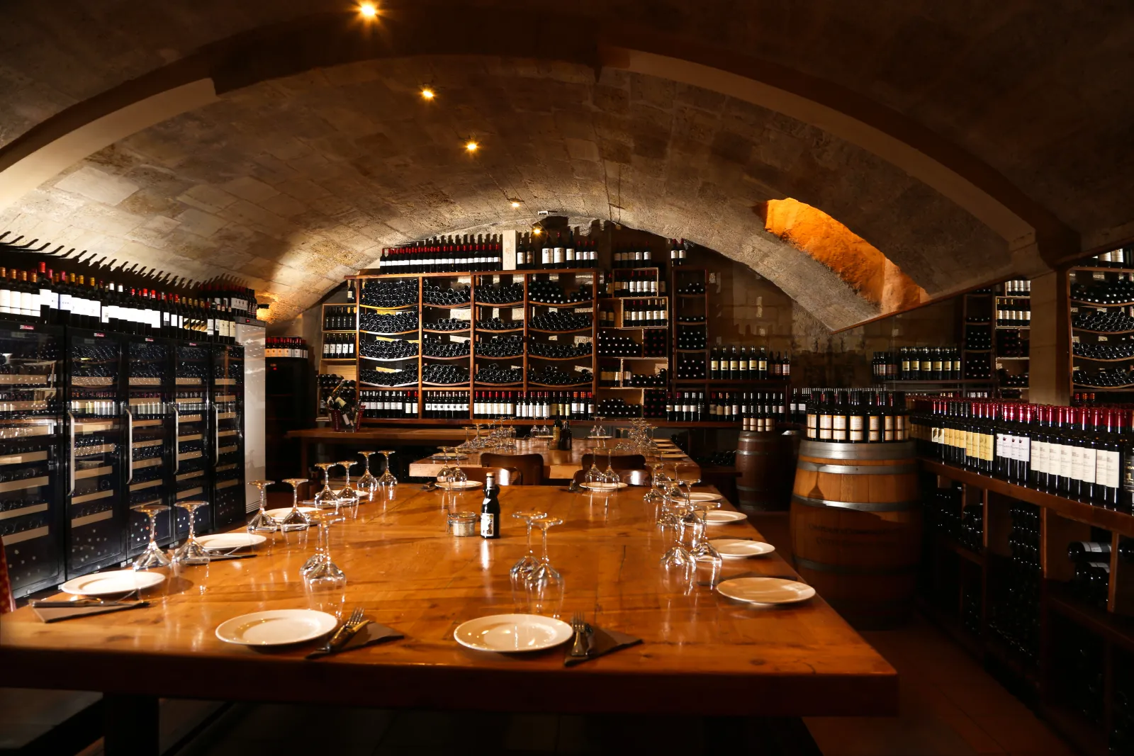 Arched underground wine cellar featuring a wooden tasting table, warm ambient lighting, and shelves stocked with Bordeaux wine bottles