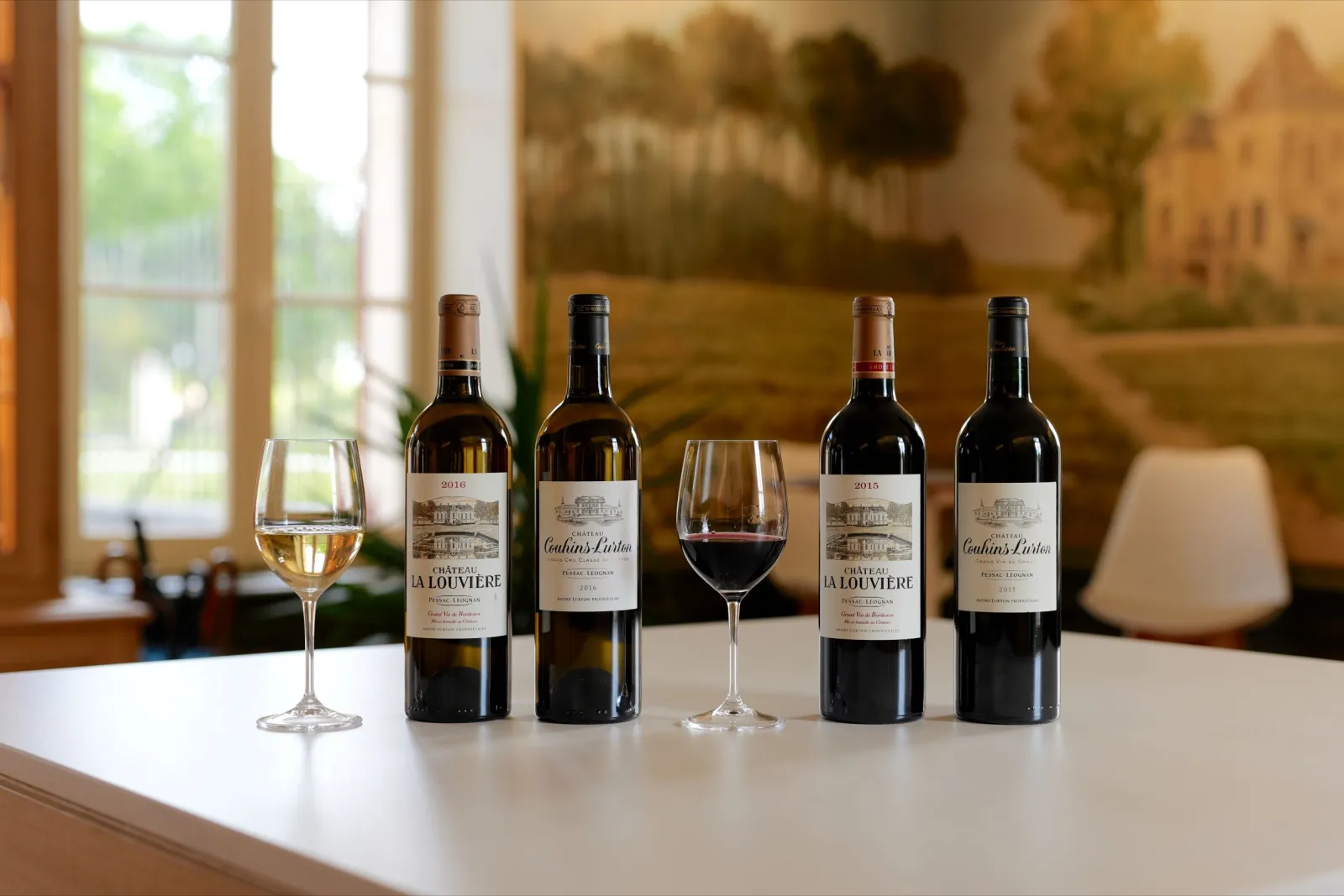 Château La Louvière wine collection from Bordeaux displayed on a white table with tasting glasses filled with white and red wine, windows and scenic landscape in the background.