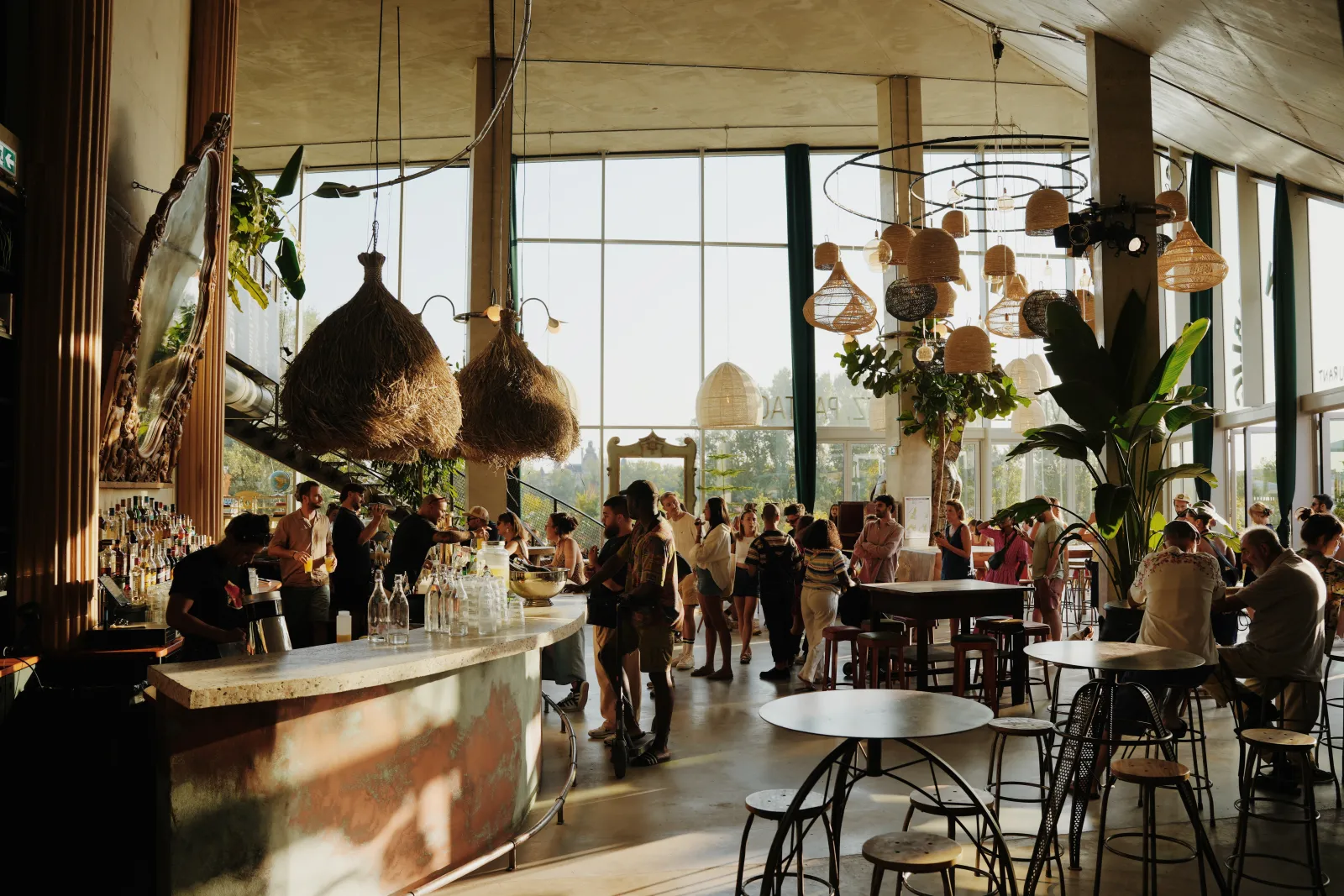 Bright interior of a modern bar in Bordeaux featuring floor-to-ceiling windows, standing and seated patrons at the counter, suspended wicker basket decorations, green plants, and a panoramic city view.