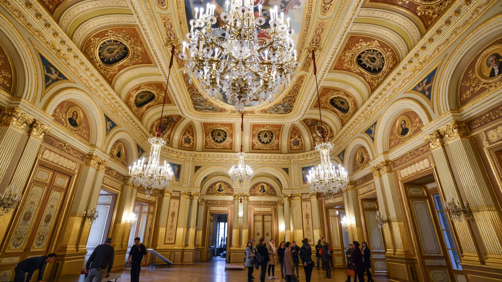 Interior of the Grand Théâtre in Bordeaux featuring ornate gilt ceiling, frescoes, crystal chandeliers, and Doric columns, with visitors admiring the baroque architecture.
