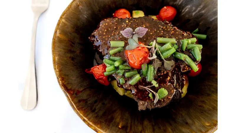 Gourmet dish plated in a glossy black bowl: braised meat topped with fresh green beans, red peppers and microgreens, served with a white fork on the side