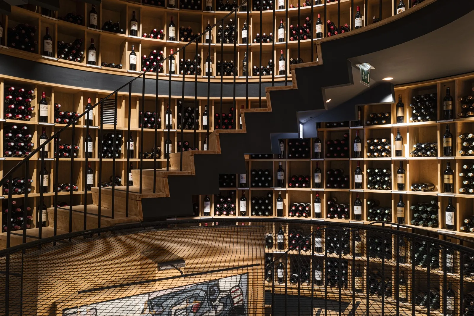 Modern circular wine cellar with illuminated wooden shelves filled with wine bottles, metal stairs in the center, and tiled flooring in contemporary architecture.