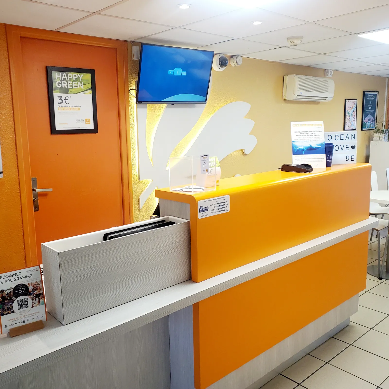 Reception desk of Hôtel Première Classe Bordeaux Sud featuring a bright orange counter, orange door, promotional Happy Green display, wall-mounted TV screen, and yellow and white wall decoration