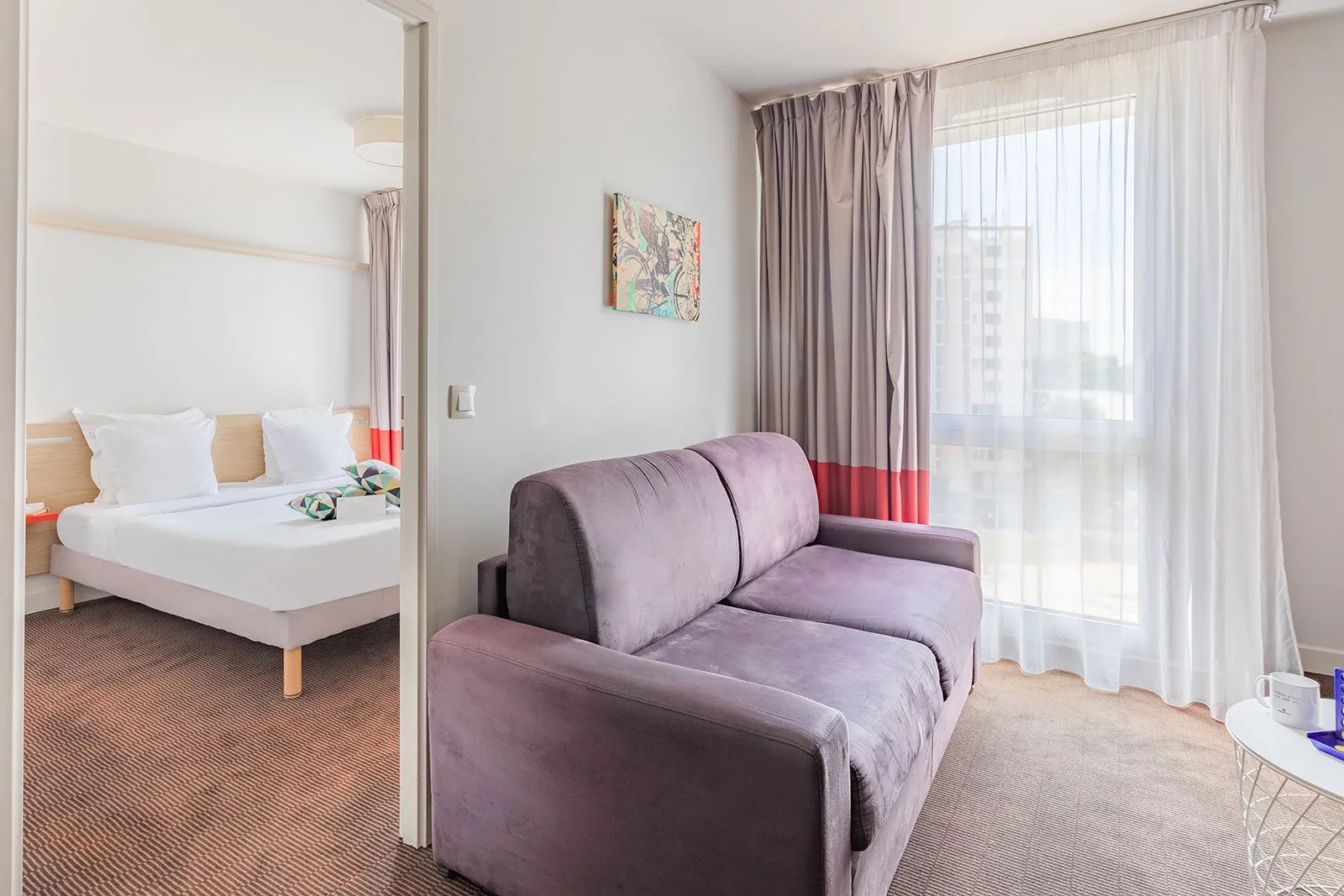 Spacious apartment room featuring a queen-size bed visible through a glass door, a purple sofa in the foreground, gray and beige curtains, and a large window overlooking Bordeaux city.
