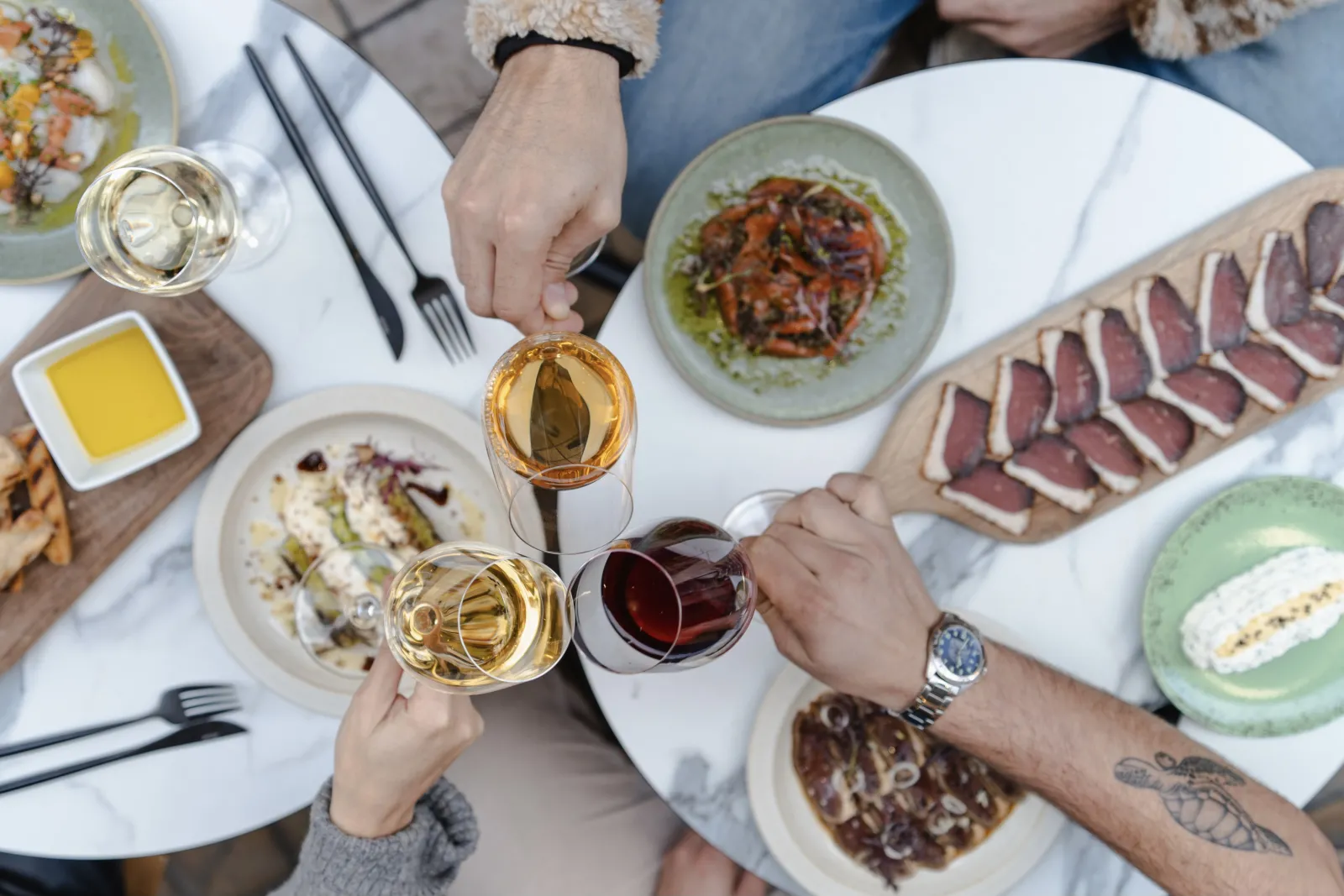 Overhead view of a gourmet tasting table in Bordeaux featuring elegant plated dishes, white and red wine glasses, cured meats, and diners toasting together.