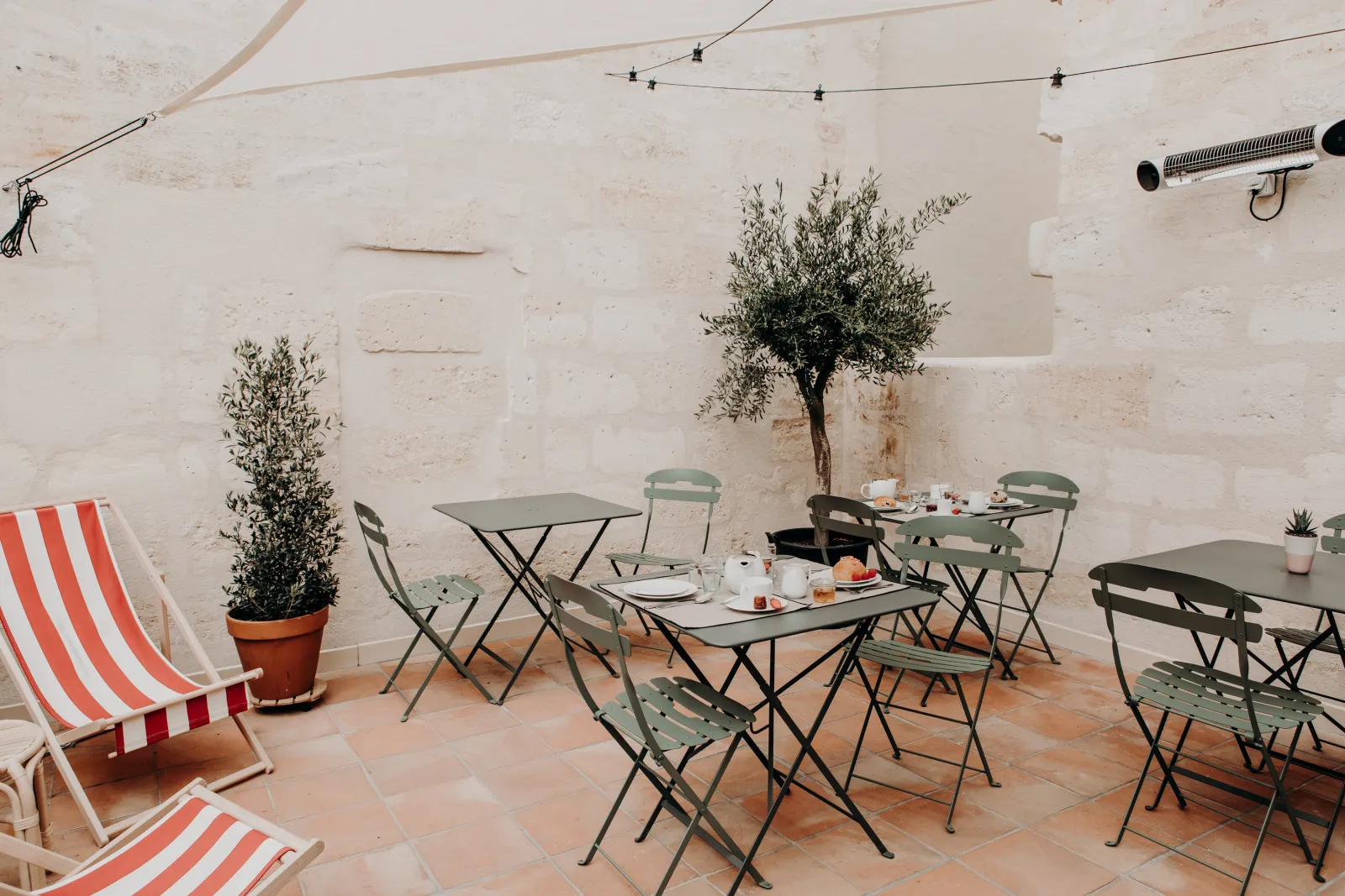 Outdoor terrace with green folding tables and chairs, potted plants, and red and white striped deck chairs at La Maison Fernand in Bordeaux.