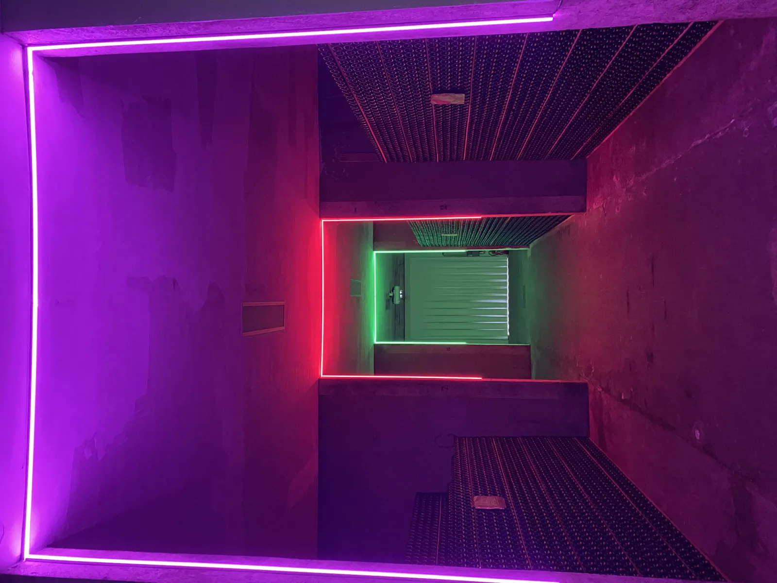 Modern interior of Cave La Rose Pauillac in Bordeaux featuring pink and magenta neon frames around a passage, window with green blinds, and metallic grid ceiling.