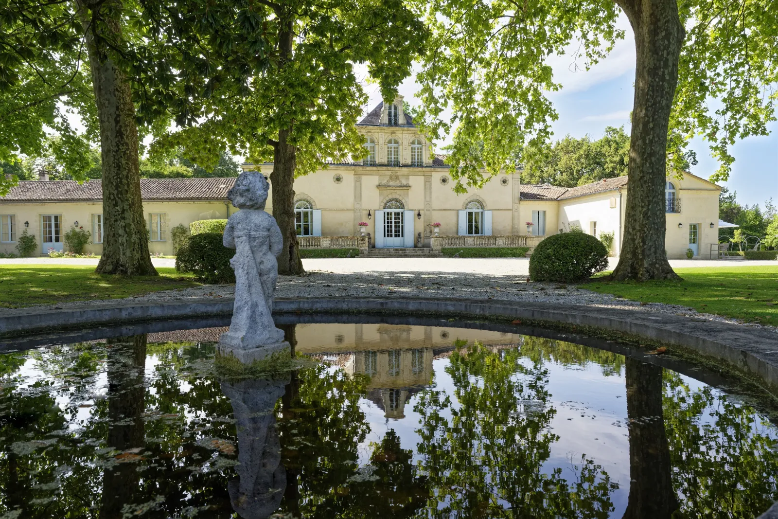Château Siran in Bordeaux reflected in a pond, surrounded by ancient trees with a stone statue in the foreground