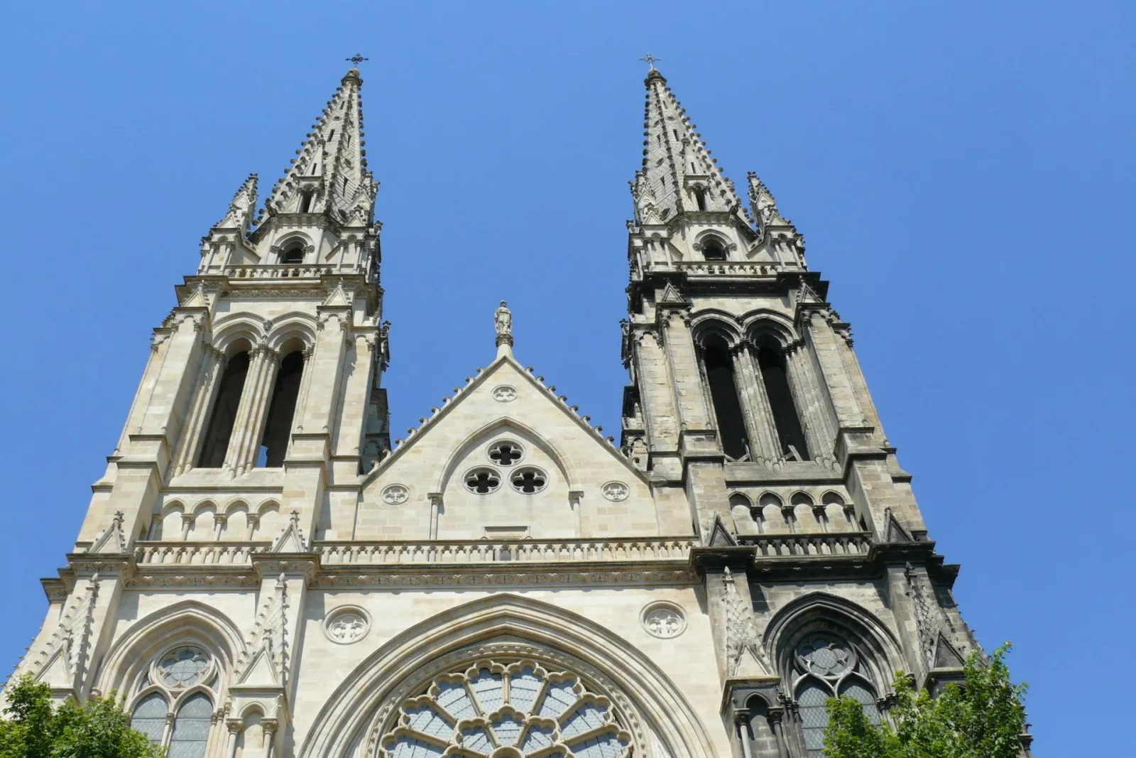 Front façade of Saint-Louis Church in Bordeaux featuring twin Gothic spires with pointed arches, a central rose window, and detailed stone architectural elements against a clear blue sky.