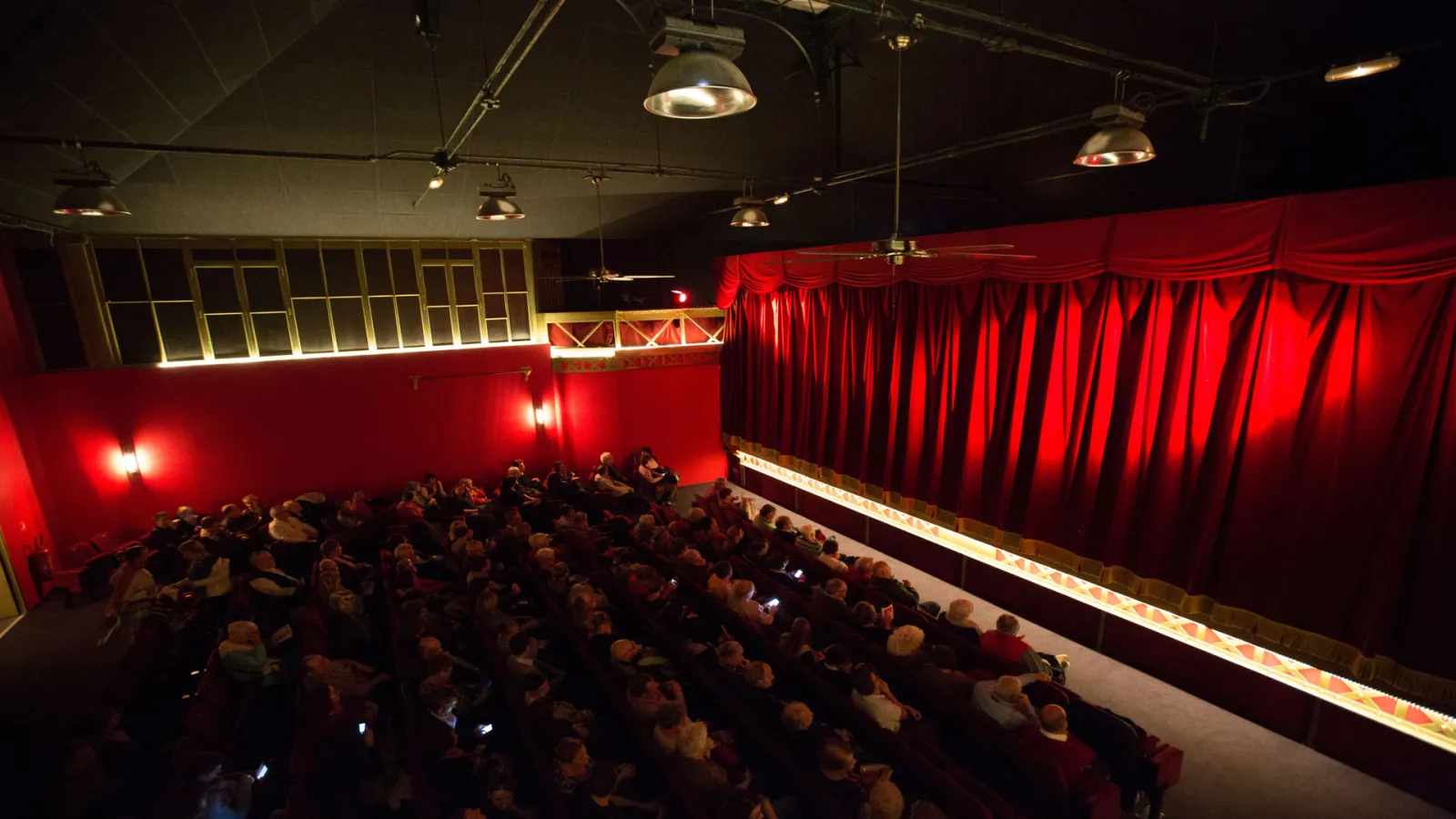 Interior of Théâtre des Salinières in Bordeaux showing a packed auditorium with spectators seated facing a stage with red curtain, dramatic red and white lighting, and suspended ceiling lights.