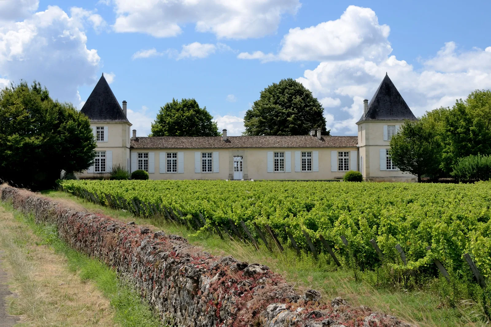 Château Climens, an elegant white château with two black conical towers, surrounded by Bordeaux vineyards under a blue cloudy sky.