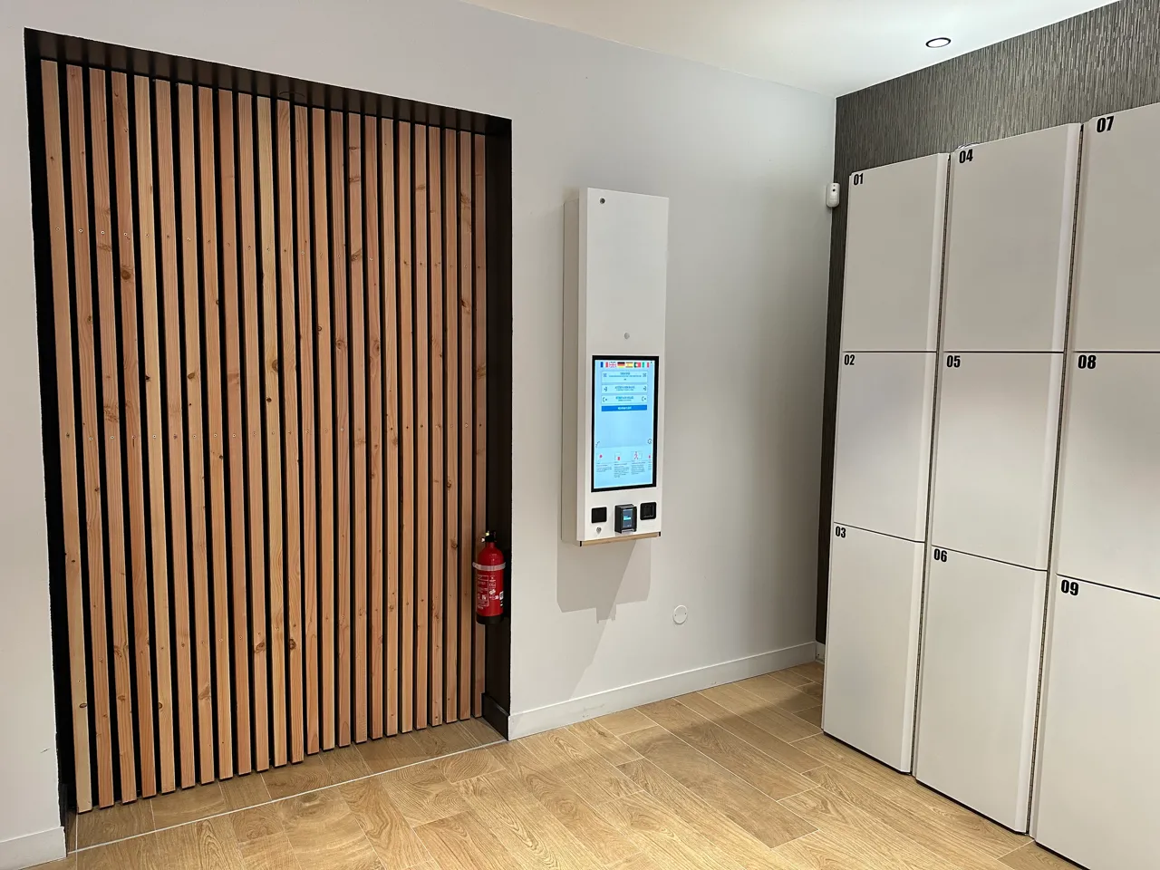 Interior of a Popin Locker automated locker service in Bordeaux featuring vertical slat wooden doors, digital management terminal, numbered white lockers and fire extinguisher.