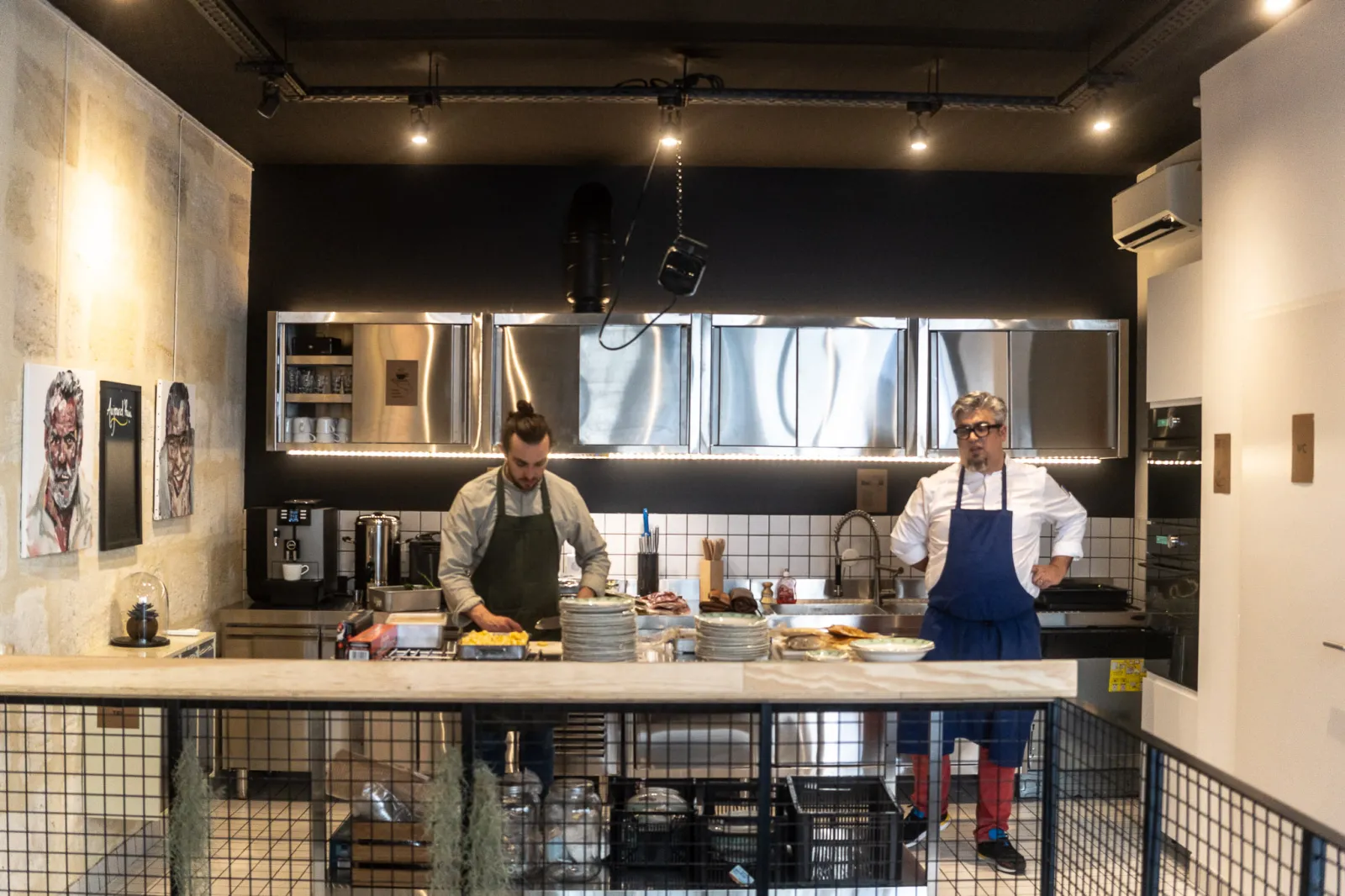 Two chefs work in a modern open kitchen at La Cuisine de Camille in Bordeaux, featuring illuminated display cases and cooking equipment.